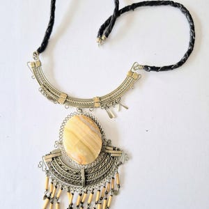 May include: A silver-toned necklace with a large, oval, light-colored stone pendant. The pendant is adorned with a fringe of small wooden beads and silver wire detailing. The necklace has a black braided cord.