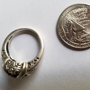 May include: A silver ring with an ornate design, featuring a central decorative element and filigree detailing. A U.S. quarter coin is next to the ring, showing the Idaho state design. The ring is on a white surface.