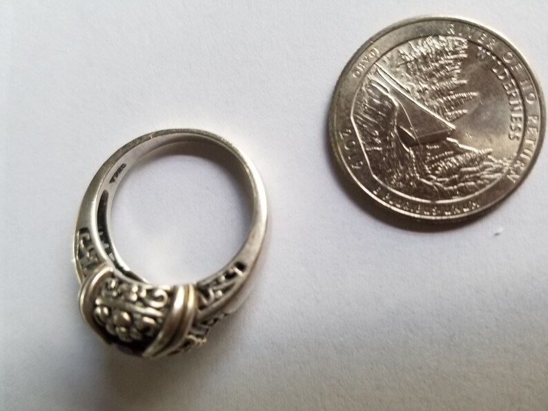 May include: A silver ring with an ornate design, featuring a central decorative element and filigree detailing. A U.S. quarter coin is next to the ring, showing the Idaho state design. The ring is on a white surface.