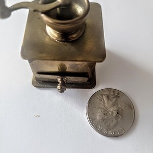 May include: A small, antique brass coffee grinder with a hand crank. The grinder is about the size of a UK 20 pence coin.