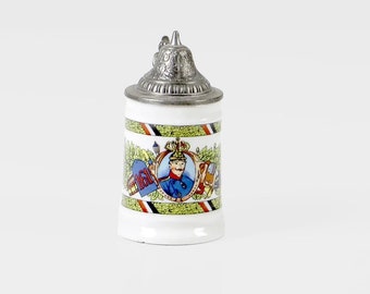 May include: A white ceramic beer stein with a silver lid. The stein features a colourful illustration of a man in a blue uniform holding a flag and a beer stein. The illustration is surrounded by a green wreath and a red, white and black ribbon.