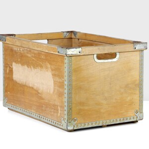May include: A wooden storage crate with metal accents. The crate has a rectangular shape and is made of natural wood with a light brown finish. The metal accents are silver and are used to reinforce the corners and edges of the crate.