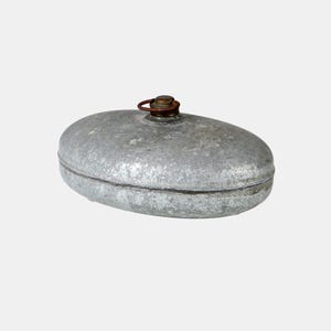 May include: Oval-shaped, vintage metal hot water bottle. The bottle is a dull silver colour with a rusty brown metal cap and handle. The bottle has a seam around the middle and is set against a plain white background.