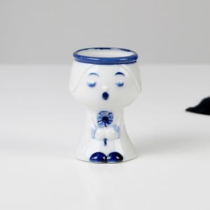 May include: A white ceramic egg cup with a blue and white design. The egg cup is shaped like a person holding a flower.