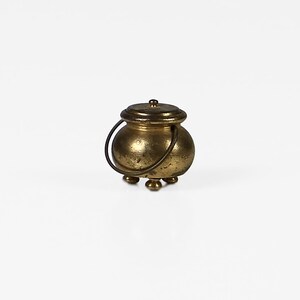 May include: A small gold cauldron with a lid and a handle. The cauldron has three feet and is made of metal.