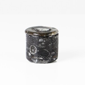 May include: A small, cylindrical container with a lid. The container is dark gray with white and light gray markings. The lid is a darker gray with a circular indentation. The container is likely made of stone or a similar material.
