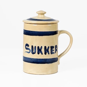 Peut inclure: Un pot à sucre en céramique blanche avec un couvercle bleu et des rayures bleues. Le pot porte l'inscription "SUKKER" en bleu.