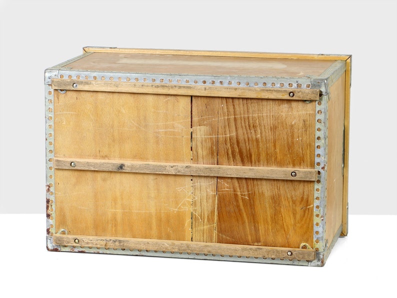 May include: A wooden storage box with metal trim and two wooden slats across the front. The box is made of light wood and has a worn, vintage look.