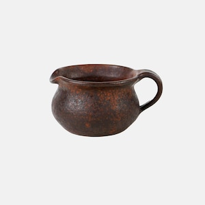 May include: A small, round ceramic creamer with a dark brown, mottled, and textured glaze. The vessel features a wide, open top with a distinct pouring spout on one side and a curved handle on the opposite side, showcasing a rustic finish.