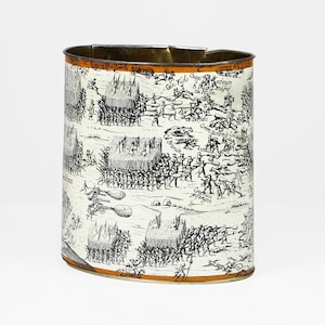 Puede incluir: Una lata de metal de forma ovalada con una ilustración en blanco y negro de una escena de batalla. La lata tiene un borde naranja.