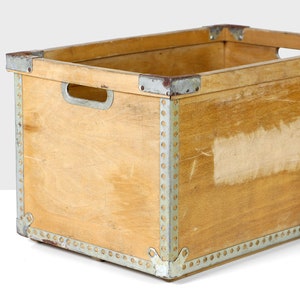 May include: A wooden storage crate with metal accents. The crate is made of light brown wood and has a rectangular shape. The metal accents are silver and are located around the edges of the crate.