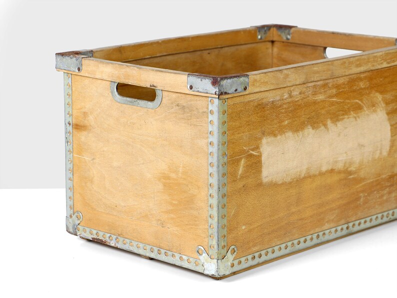 May include: A wooden storage crate with metal accents. The crate is made of light brown wood and has a rectangular shape. The metal accents are silver and are located around the edges of the crate.