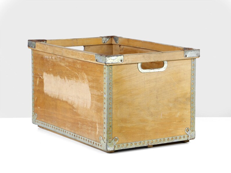 May include: A wooden storage crate with metal accents. The crate has a rectangular shape and is made of natural wood with a light brown finish. The metal accents are silver and are used to reinforce the corners and edges of the crate.