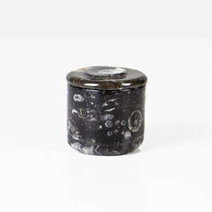 May include: A small, cylindrical container with a lid. The container is primarily black with white and gray marbling. The lid is a darker shade of black with a circular, lighter-colored design in the center. The container is likely made of stone.