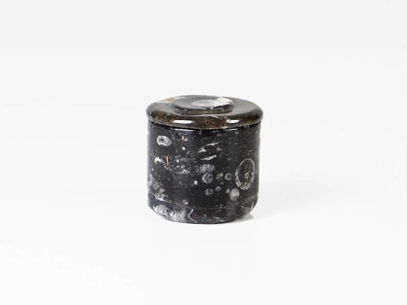 May include: A small, cylindrical container with a lid. The container is primarily black with white and gray marbling. The lid is a darker shade of black with a circular, lighter-colored design in the center. The container is likely made of stone.