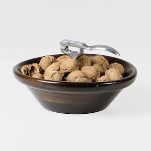 Könnte beinhalten: Eine dunkelbraune Schüssel gefüllt mit Walnüssen und einem silbernen Nussknacker. Die Schüssel hat einen breiten Rand und eine glatte Oberfläche. Die Walnüsse sind braun und haben strukturierte Schalen. Der Nussknacker ist über den Walnüssen positioniert.