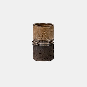 Può includere: Un vaso cilindrico in ceramica con superficie strutturata. La metà superiore è marrone chiaro, la metà inferiore è marrone scuro. Una fascia di materiale strutturato separa le due metà. Il vaso ha un design semplice e minimalista.