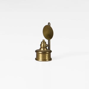 Pode incluir: Uma pequena lamparina de óleo de latão antigo com base redonda e abajur circular. A lamparina tem um design detalhado e uma estética vintage, ideal para colecionadores. O fundo é branco.