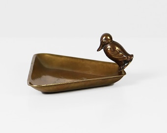 May include: A bronze-colored, rectangular tray with a duck figurine on one end. The tray has a slightly angled design and a smooth, reflective surface. The duck is detailed with a curved neck and a small beak.