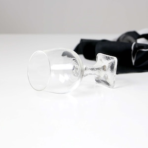 May include: A clear glass wine goblet with a textured, square-shaped stem. The goblet is lying on its side, with a black and white fabric in the background. The glass is empty and reflects the light.