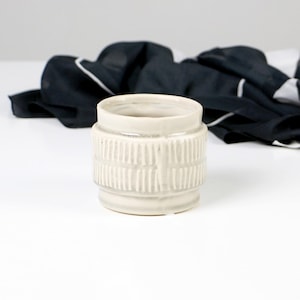 Könnte beinhalten: Ein weißer Keramiktopf mit einem strukturierten Design. Der Topf hat eine zylindrische Form und eine glatte, weiße Glasur.