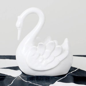 Peut inclure: Une figurine de cygne en céramique blanche brillante. Le cygne a un cou courbé et des plumes d'ailes détaillées. La figurine est posée sur une surface à motifs noirs et blancs, créant un contraste saisissant. Une pièce décorative élégante.