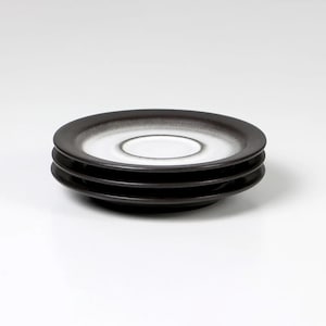 Könnte beinhalten: Drei schwarze und weiße Keramikuntertassen, die übereinander gestapelt sind. Die Untertassen haben eine weiße Mitte und einen schwarzen Rand.