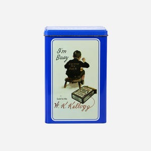 Op de afbeelding: Een rechthoekige blauwe blikken doos met een wit etiket. Het etiket toont een vintage illustratie van een jongen op een kruk. Tekst op de doos luidt "I'm Busy, Excuse My Back" en "W. K. Kellogg".