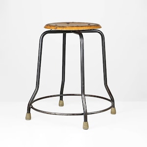 Könnte beinhalten: Ein Hocker im Vintage-Stil mit einer runden, hellbraunen Holzsitze und einem dunklen Metallrahmen. Der Rahmen hat vier gebogene Beine, die durch eine kreisförmige Basis verbunden sind, mit Gummifüßen. Der Hocker ist zum Sitzen konzipiert.