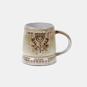 Peut inclure: Une tasse en céramique avec une finition émaillée réactive beige et brune. La tasse présente un motif floral en creux sur le devant, entouré d'une bande de petites indentations circulaires près des bords supérieur et inférieur. Anse incurvée.
