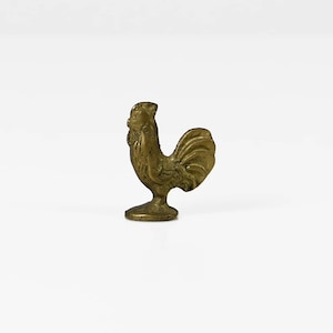 May include: A small, gold-colored metal rooster figurine with a detailed design. The rooster is standing on a small base.