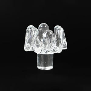 May include: A clear glass decorative object with an unusual, organic form. It has a cylindrical base supporting a cluster of rounded, petal-like shapes. The transparent glass allows light to pass through, set against a black background.