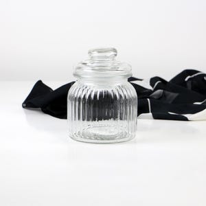 May include: A clear glass jar with a lid. The jar has vertical ribbed detailing and a rounded shape. A black and white patterned cloth is in the background.