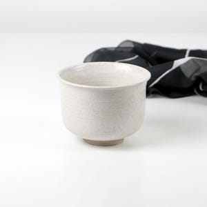Könnte beinhalten: Eine kleine, runde, cremeweiße Keramikschale mit gesprenkelter Textur und einem glatten, leicht ausgestellten Rand. Die Schale hat einen dicken, stabilen Boden und eine matte Oberfläche und steht vor einem gefalteten schwarzen Stoff mit weißen Linien.