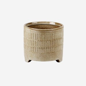 May include: A beige ceramic planter with a cylindrical shape and a textured, vertical striped pattern. The planter has three small feet and a neutral color, suitable for various plants. The interior is a darker shade of brown.