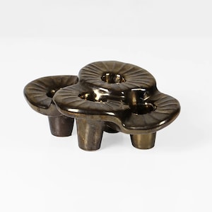 May include: A four-armed, three-candle holder made of ceramic with a dark brown glaze. The holder has a unique, organic shape with rounded edges and a textured surface.