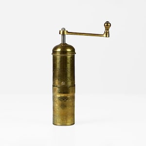 Puede incluir: Un molinillo de pimienta antiguo de latón con cuerpo cilíndrico y manivela. El molinillo tiene un aspecto envejecido, con patrones decorativos grabados alrededor del cuerpo. El mango y la parte superior también son de latón, sobre fondo blanco.