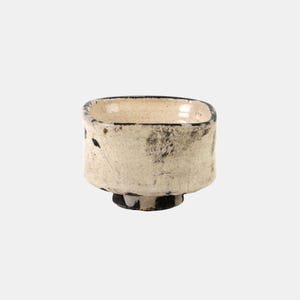 May include: A small, square-shaped ceramic bowl with rounded corners, featuring a cream-coloured glaze with dark, mottled charcoal-coloured markings and a visible crackle texture. The bowl sits on a short, dark-coloured circular pedestal base.