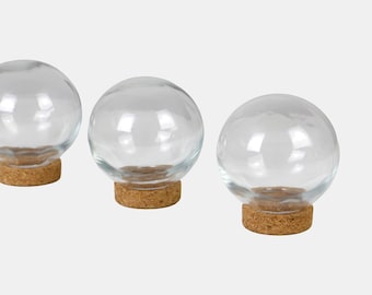 Op de afbeelding: Drie heldere glazen bolvormige containers met kurken bodems. De glazen bollen zijn transparant, waardoor je er doorheen kunt kijken. De kurken bodems zijn lichtbruin van kleur. De containers zijn in een rij geplaatst tegen een witte achtergrond.
