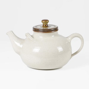May include: A white ceramic teapot with a brown lid and a brown knob. The teapot has a rounded body and a spout on the side.