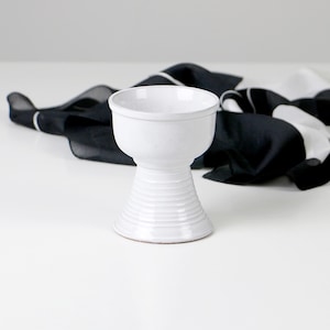 Könnte beinhalten: Ein weißer Keramikkelch mit geripptem Fuß und breiter, offener Oberseite. Der Kelch steht vor einem Hintergrund aus schwarz-weiß gemustertem Stoff. Das Design ist schlicht und elegant, geeignet für dekorative Zwecke.