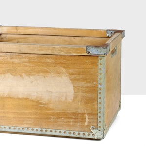 May include: A wooden crate with metal accents. The crate is made of light brown wood and has a worn, rustic look. The metal accents are silver and are used to reinforce the corners and edges of the crate.