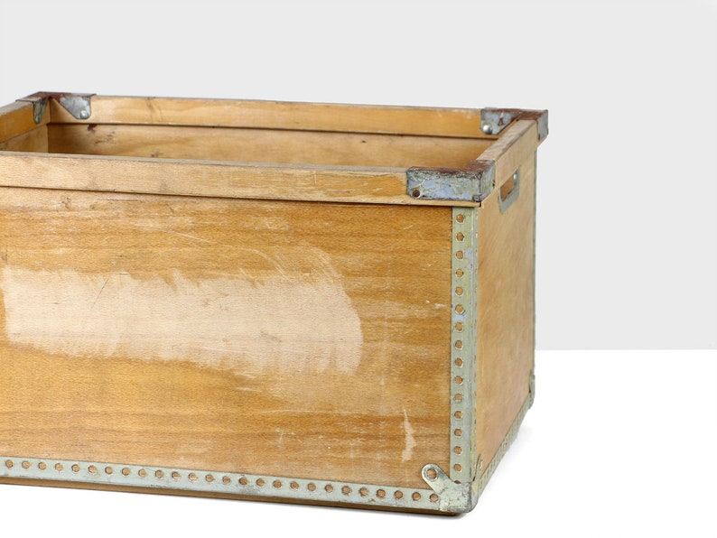 May include: A wooden crate with metal accents. The crate is made of light brown wood and has a worn, rustic look. The metal accents are silver and are used to reinforce the corners and edges of the crate.