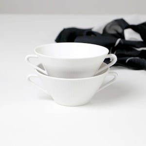 Op de afbeelding: Twee witte keramische soepkommen op elkaar gestapeld. Elke kom heeft een handvat en een geribbelde textuur. Een zwart-wit patroon sjaal staat op de achtergrond.