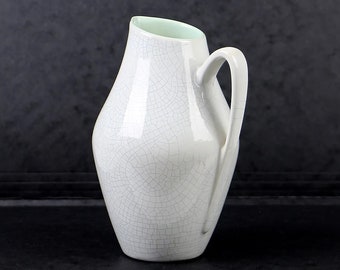 May include: A white ceramic pitcher with a light green rim and a crackled glaze. The pitcher has a rounded body and a single handle.