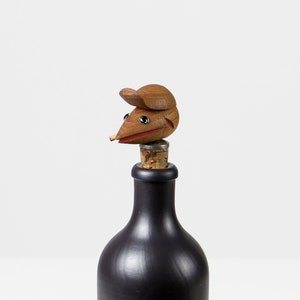 May include: A wooden bottle stopper shaped like a mouse with a red mouth and green eyes. The stopper is on top of a dark brown glass bottle.