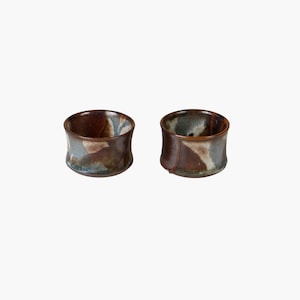 May include: Two small ceramic bowls with a brown, blue, and white glaze. The bowls are slightly flared at the top and have a rounded bottom.