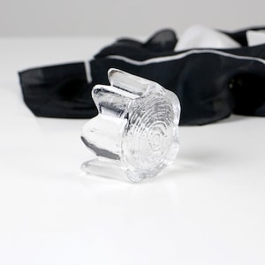 May include: A clear glass candle holder with a textured, tree-ring design. The candle holder has a unique shape with three protruding sections. A black and white scarf is in the background.