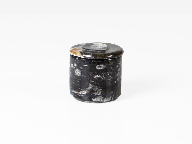 May include: A small, cylindrical container with a lid. The container is made of a dark, speckled material, with a mix of black, gray, and white tones. The lid is a similar material, with a slightly lighter color and a circular shape.