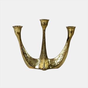 May include: A gold-coloured, three-armed candelabra with a textured surface. The candle holders are positioned at varying heights, with a central, upright holder and two curved arms extending outwards. The design is symmetrical and decorative.